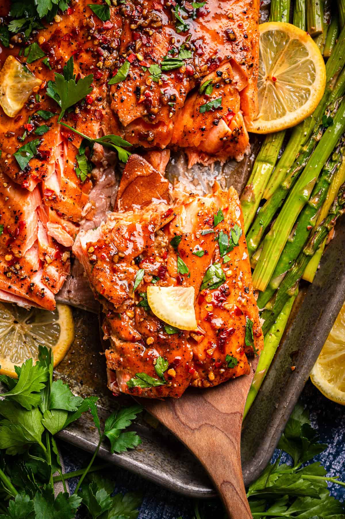 A wooden spatula lifts a piece of baked salmon topped with herbs and lemon slices from a sheet pan, surrounded by asparagus spears, fresh parsley, and more lemon slices. The salmon is glazed and garnished with seasoning.