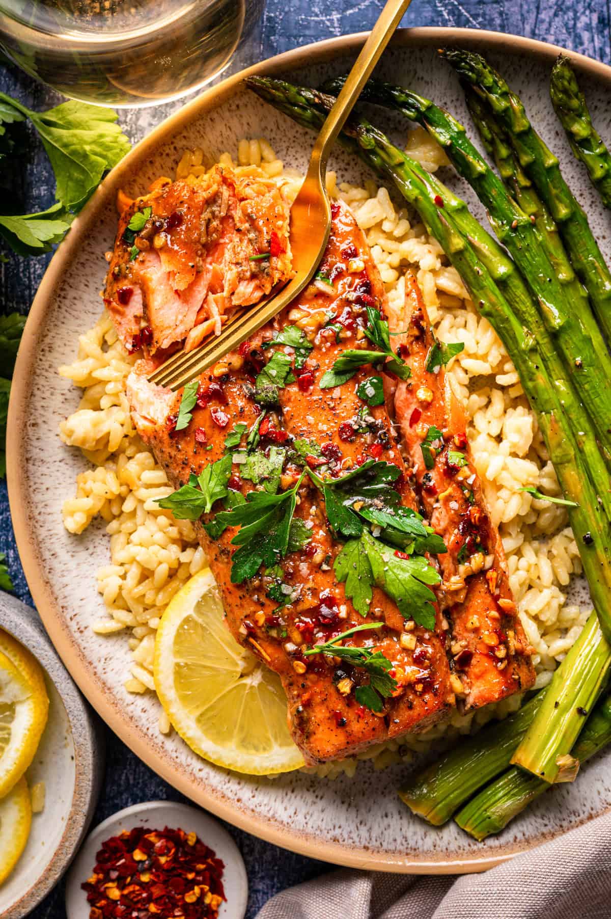 A plate of cooked salmon fillet garnished with fresh herbs, served over rice with roasted asparagus and a lemon slice. A fork is flaking the salmon, and red pepper flakes are visible on top.