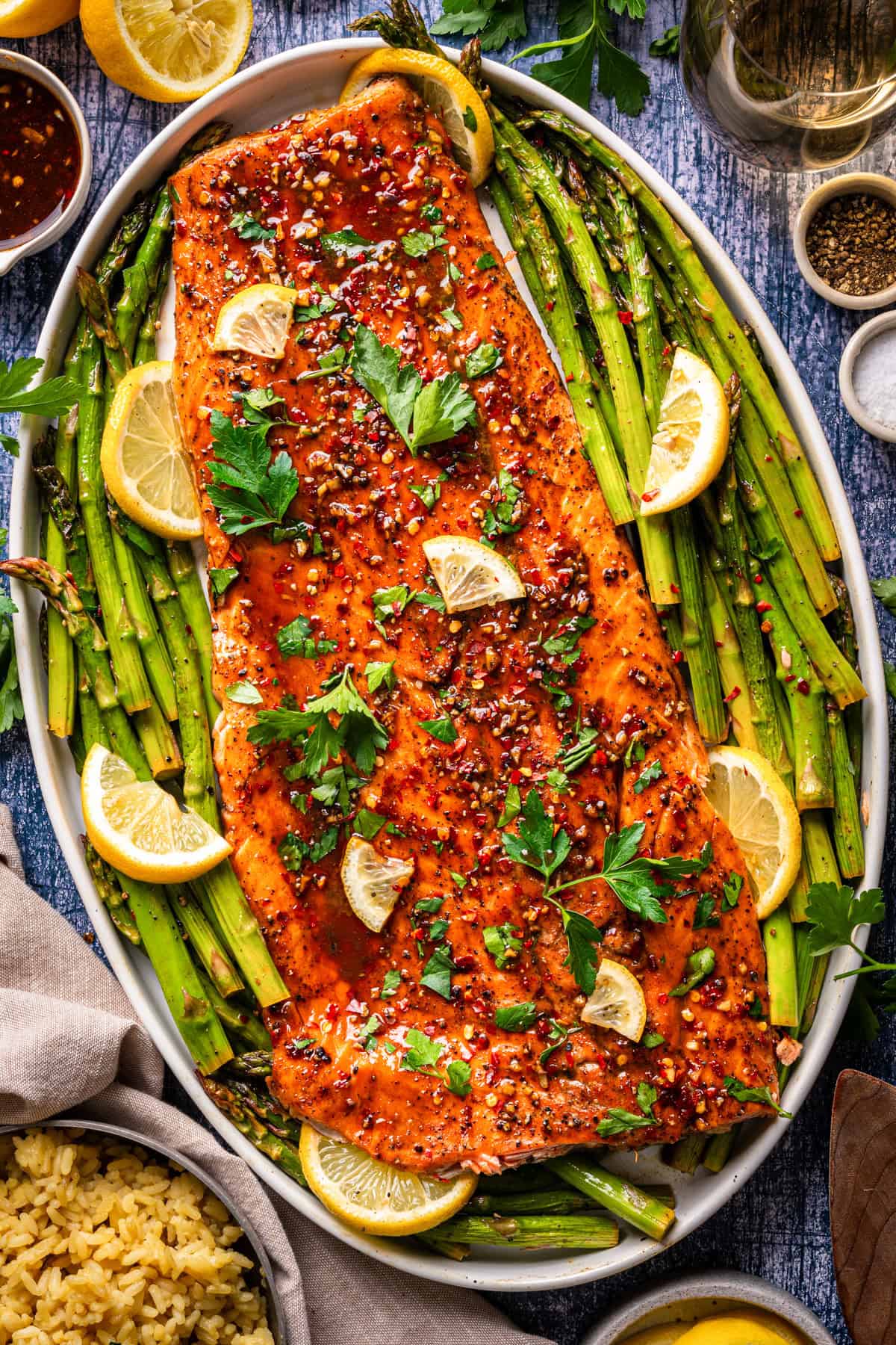 A large roasted salmon fillet garnished with lemon wedges, parsley, and chili flakes sits on a bed of asparagus on an oval platter, with rice and lemon slices nearby on a rustic table.