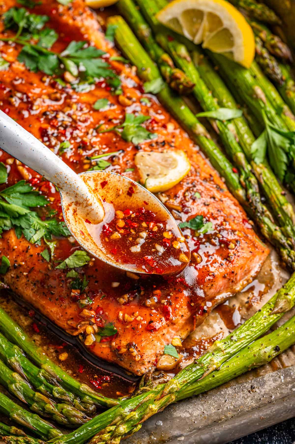 A baked salmon fillet topped with maple glaze and herbs, surrounded by roasted asparagus and lemon slices, with a spoon drizzling sauce over the salmon.