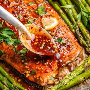 A close-up of baked salmon topped with a maple glaze, being spooned over the fish, surrounded by roasted asparagus and garnished with fresh herbs and a lemon slice.