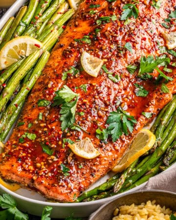 A large baked salmon fillet topped with lemon slices, parsley, and spices, served on a platter with roasted asparagus and lemon wedges, garnished with red pepper flakes.