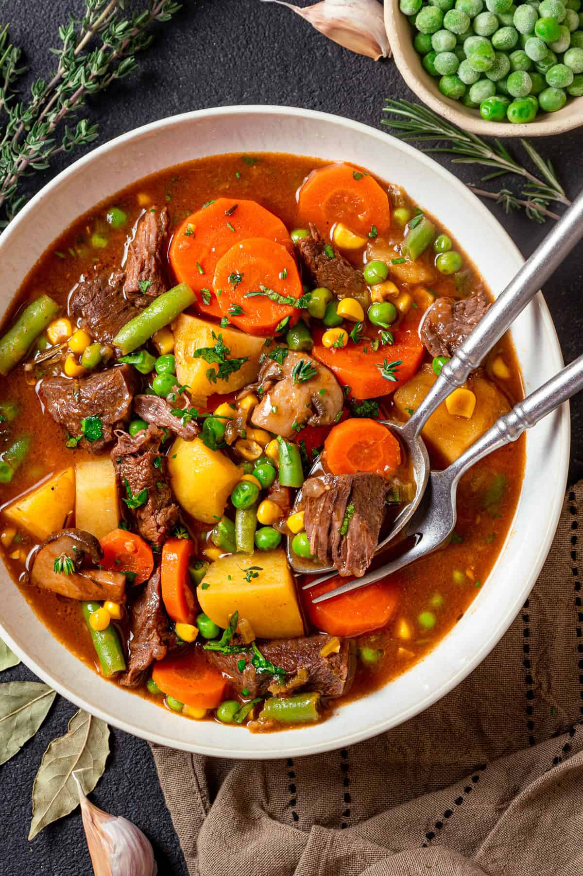 Mulligan Stew (Hobo Stew) A bowl of mulligan stew filled with chunks of beef, potatoes, carrots, green beans, peas, and corn in a rich broth, garnished with fresh herbs, with a spoon and a fork resting inside the bowl.