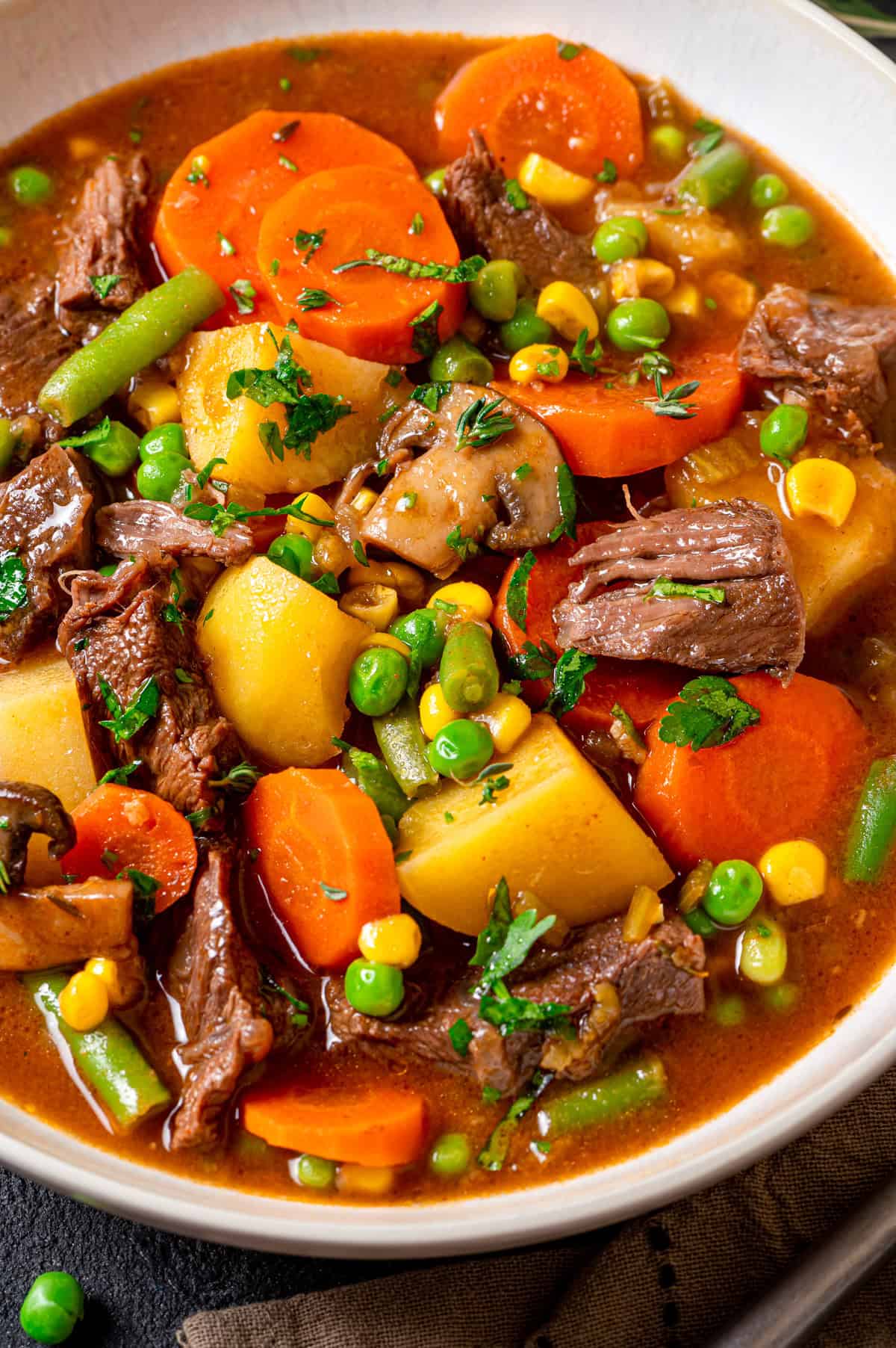 Mulligan Stew (Hobo Stew) A close-up of a hearty mulligan stew with chunks of beef, potatoes, carrots, green beans, peas, corn, and mushrooms in a rich brown broth, garnished with fresh parsley.