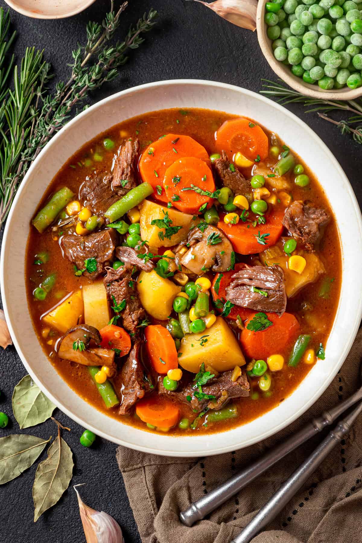 Mulligan Stew (Hobo Stew) A bowl of hearty mulligan stew filled with chunks of beef, carrots, potatoes, peas, corn, and green beans, garnished with herbs. Fresh rosemary, bay leaves, and frozen peas are visible around the bowl.