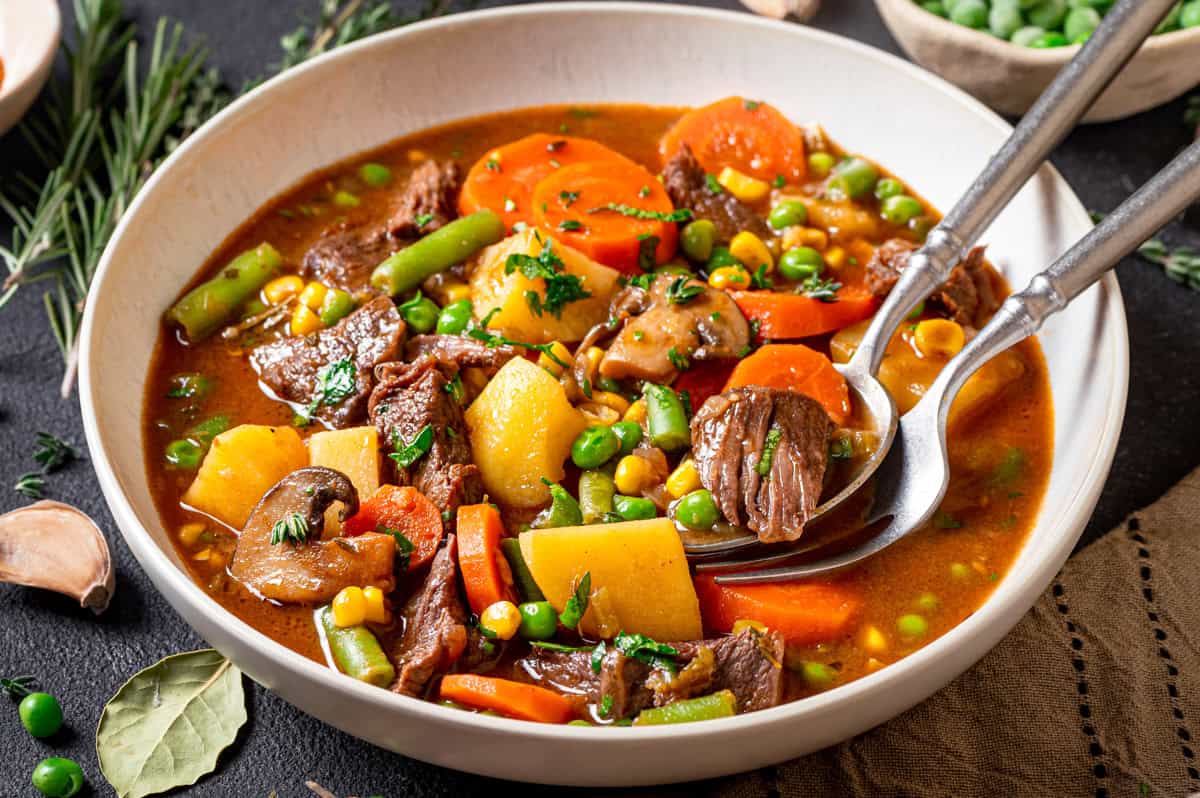 Mulligan Stew, also known as Hobo Stew, is a classic one-pot beef-and-vegetable soup featuring tender beef, potatoes, carrots, corn, peas, and a rich, savory beef broth. A bowl of hearty mulligan stew with chunks of beef, potatoes, carrots, green beans, peas, corn, and mushrooms in a rich broth, garnished with fresh herbs. A spoon and a fork rest in the bowl.