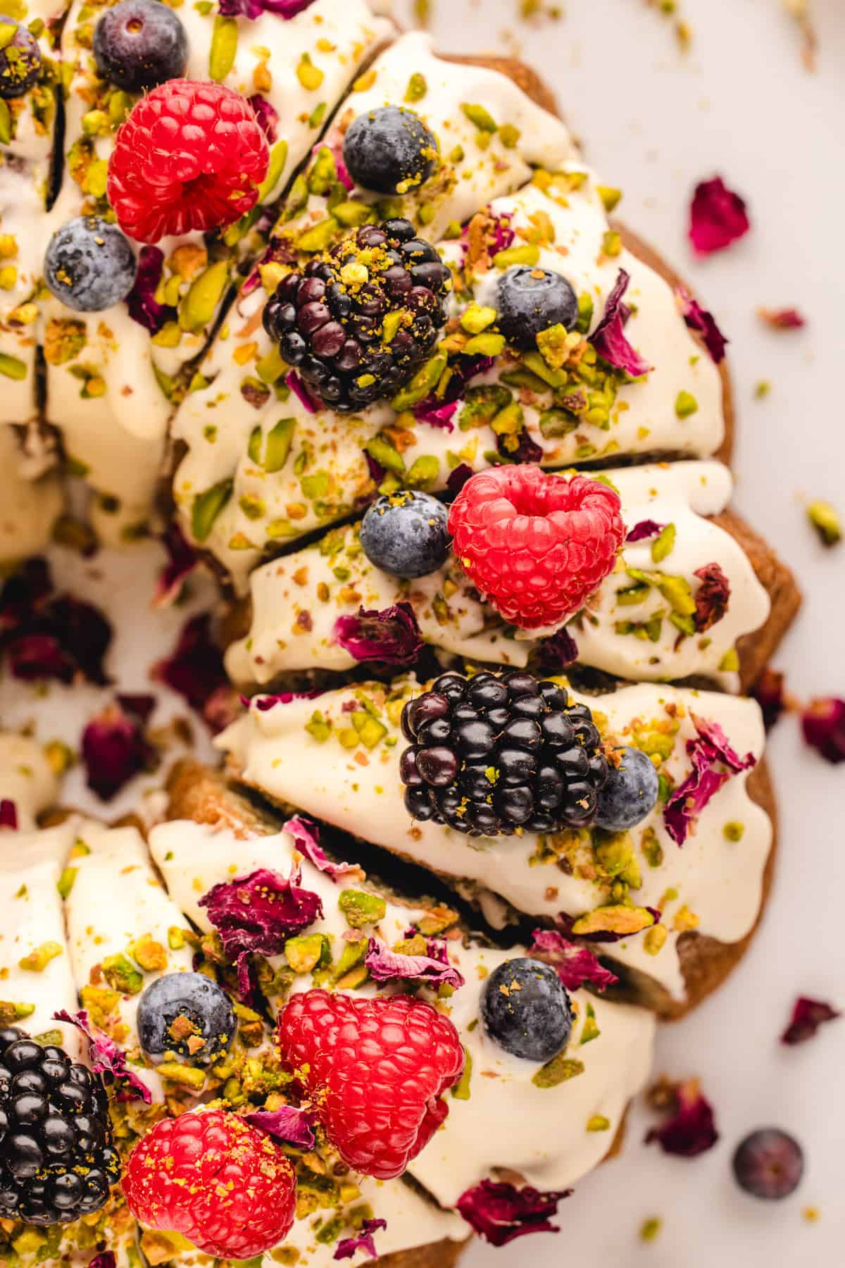 A close-up of a decorated pistachio bundt cake topped with white icing, fresh raspberries, blackberries, blueberries, chopped pistachios, and dried rose petals on a white surface.