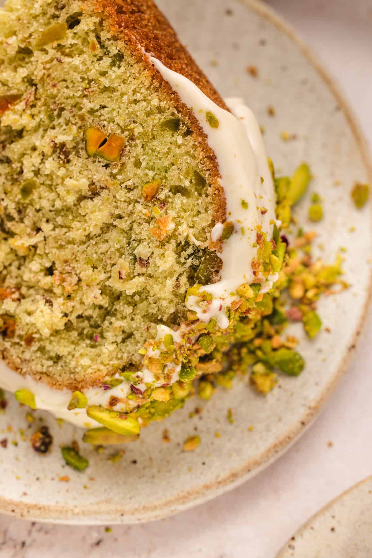 A close-up of a slice of green pistachio bundt cake with white icing, topped with chopped pistachios, on a beige ceramic plate. Crumbs and pistachio pieces are scattered around the slice.