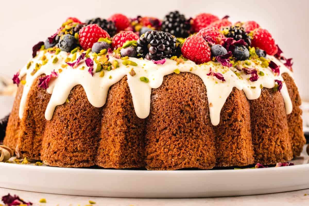 A pistachio Bundt cake topped with white icing, fresh blackberries, raspberries, blueberries, chopped pistachios, and dried flower petals, displayed on a white plate.