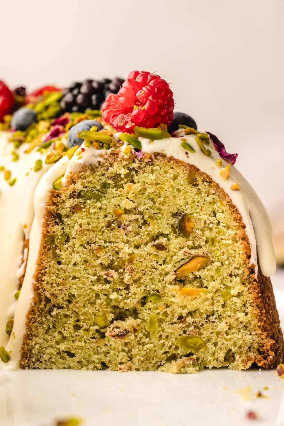 Close-up of a sliced pistachio bundt cake with a light green interior, topped with white icing, crushed pistachios, and fresh berries including raspberry, blueberry, and blackberry.