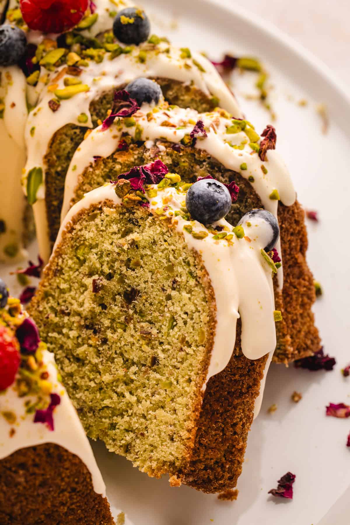 A close-up of sliced pistachio bundt cake topped with white icing, blueberries, chopped pistachios, and edible flower petals on a white plate.