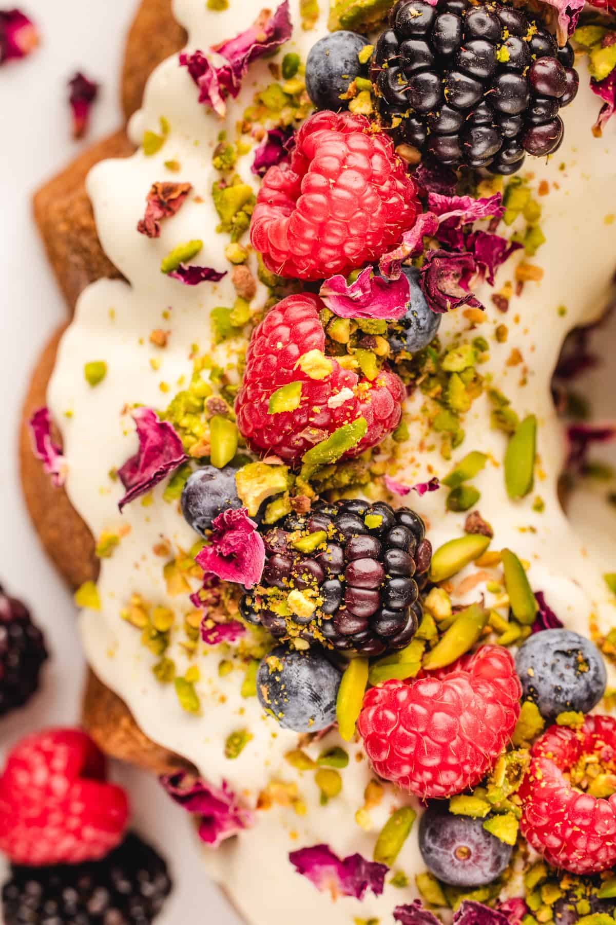 A close-up of a pistachio bundt cake topped with white icing, fresh raspberries, blackberries, blueberries, chopped pistachios, and dried rose petals. 
