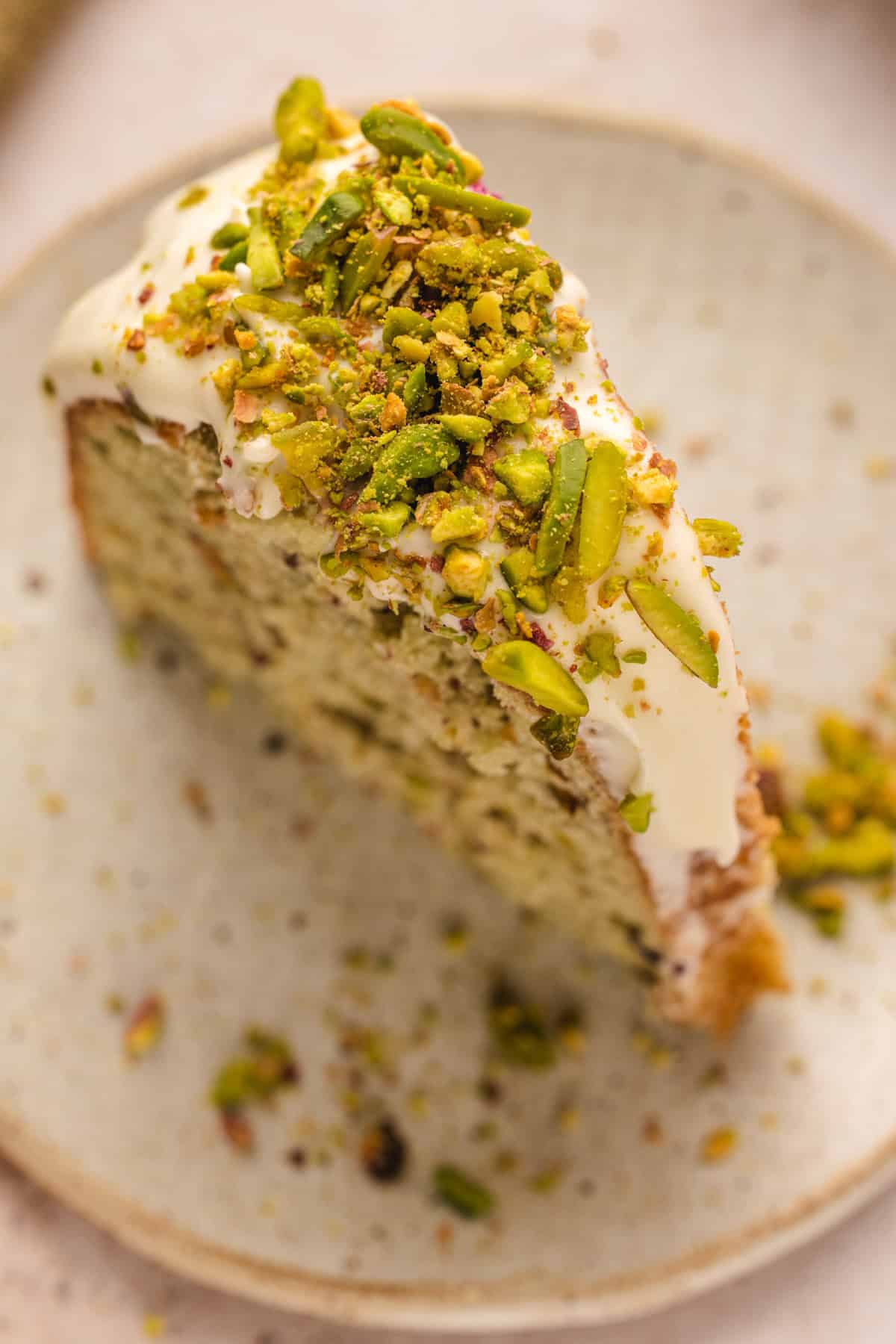 A close-up of a slice of pistachio bundt cake topped with white icing and a generous sprinkle of chopped pistachios, served on a light-colored ceramic plate.