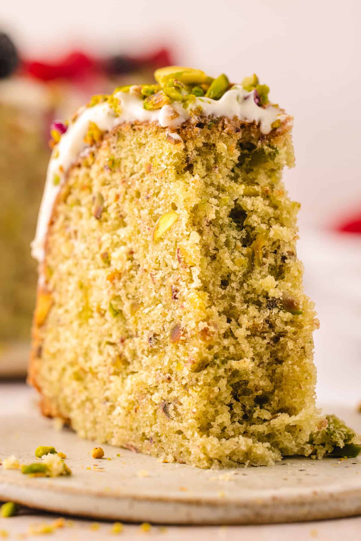 A close-up of a slice of pistachio cake topped with white icing and chopped pistachios, resting on a ceramic plate. The cake appears moist and fluffy with visible pistachio pieces inside.