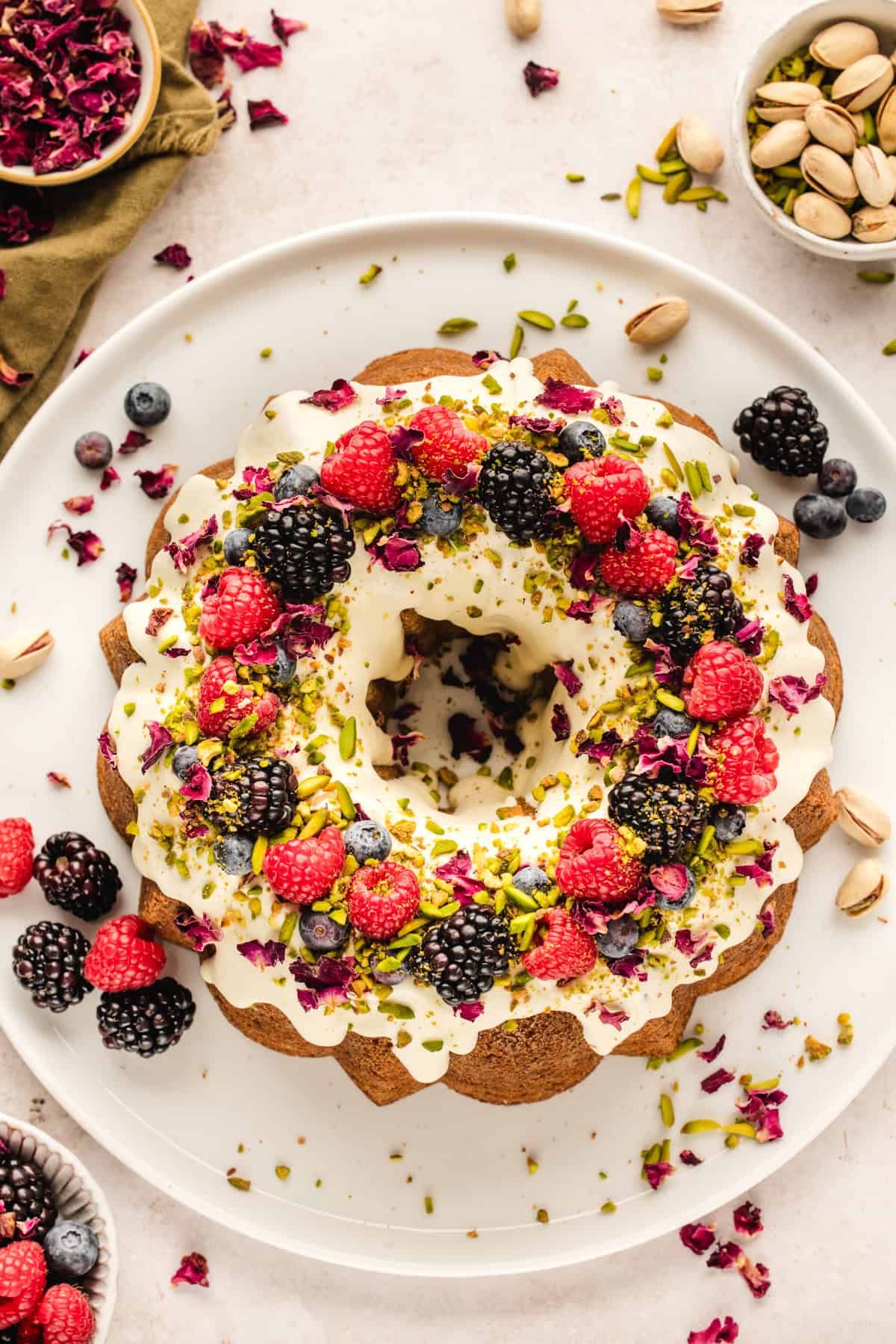 A pistachio Bundt cake topped with white icing, fresh raspberries, blackberries, blueberries, chopped pistachios, and edible flower petals, displayed on a white plate with bowls of fruit and nuts nearby.
