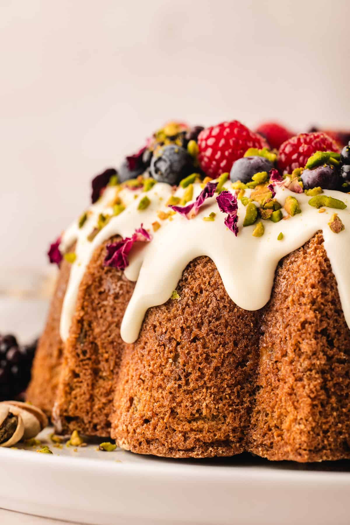 A close-up of a pistachio bundt cake topped with white icing, chopped pistachios, and assorted berries including raspberries and blueberries, displayed on a white plate.