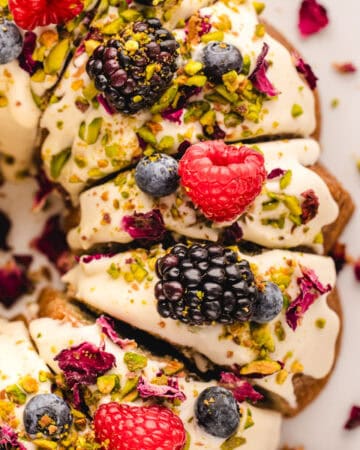 Close-up of a Pistachio Bundt Cake topped with cream, fresh raspberries, blackberries, blueberries, chopped pistachios, and dried rose petals, arranged in slices on a white surface.