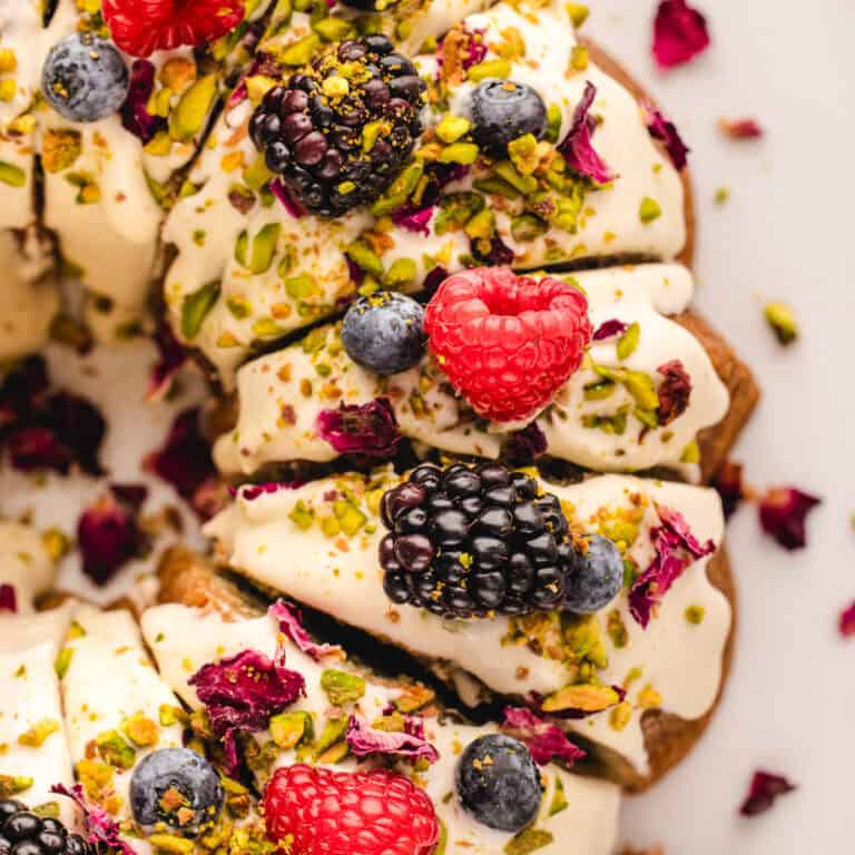 Close-up of a Pistachio Bundt Cake topped with cream, fresh raspberries, blackberries, blueberries, chopped pistachios, and dried rose petals, arranged in slices on a white surface.