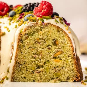 A close-up of a sliced pistachio bundt cake topped with white icing, fresh berries, chopped pistachios, and rose petals, showing the cake's green, nut-studded interior.