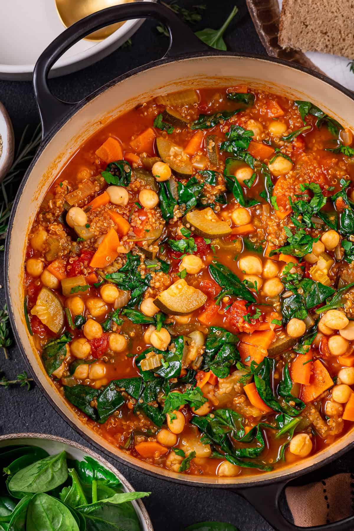 A pot filled with colorful vegetable quinoa soup featuring chickpeas, carrots, zucchini, spinach, tomatoes, and greens in a savory broth, viewed from above. A bowl of fresh spinach and a plate of bread are nearby.