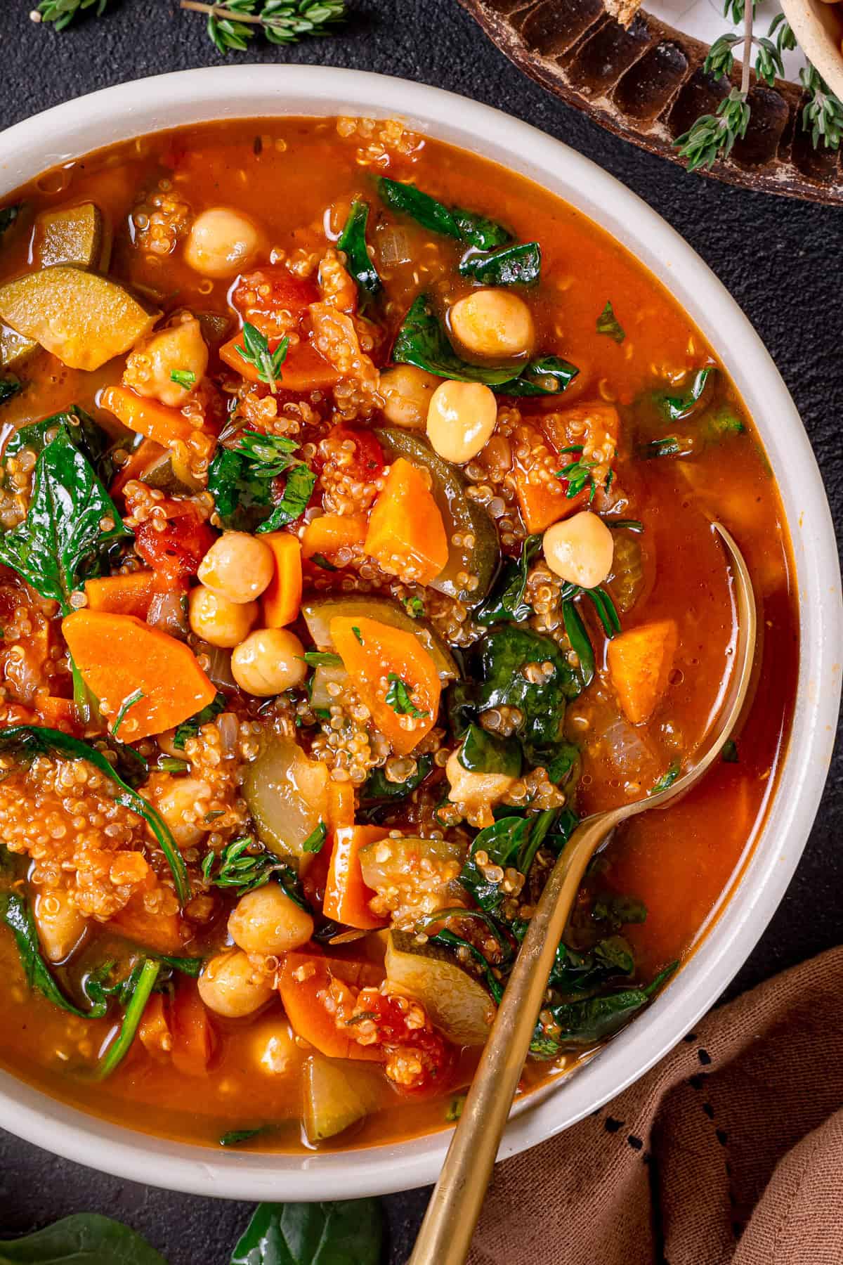 A bowl of hearty vegetable quinoa soup with chickpeas, quinoa, spinach, carrots, zucchini, and tomatoes, shown with a gold spoon on the side. 