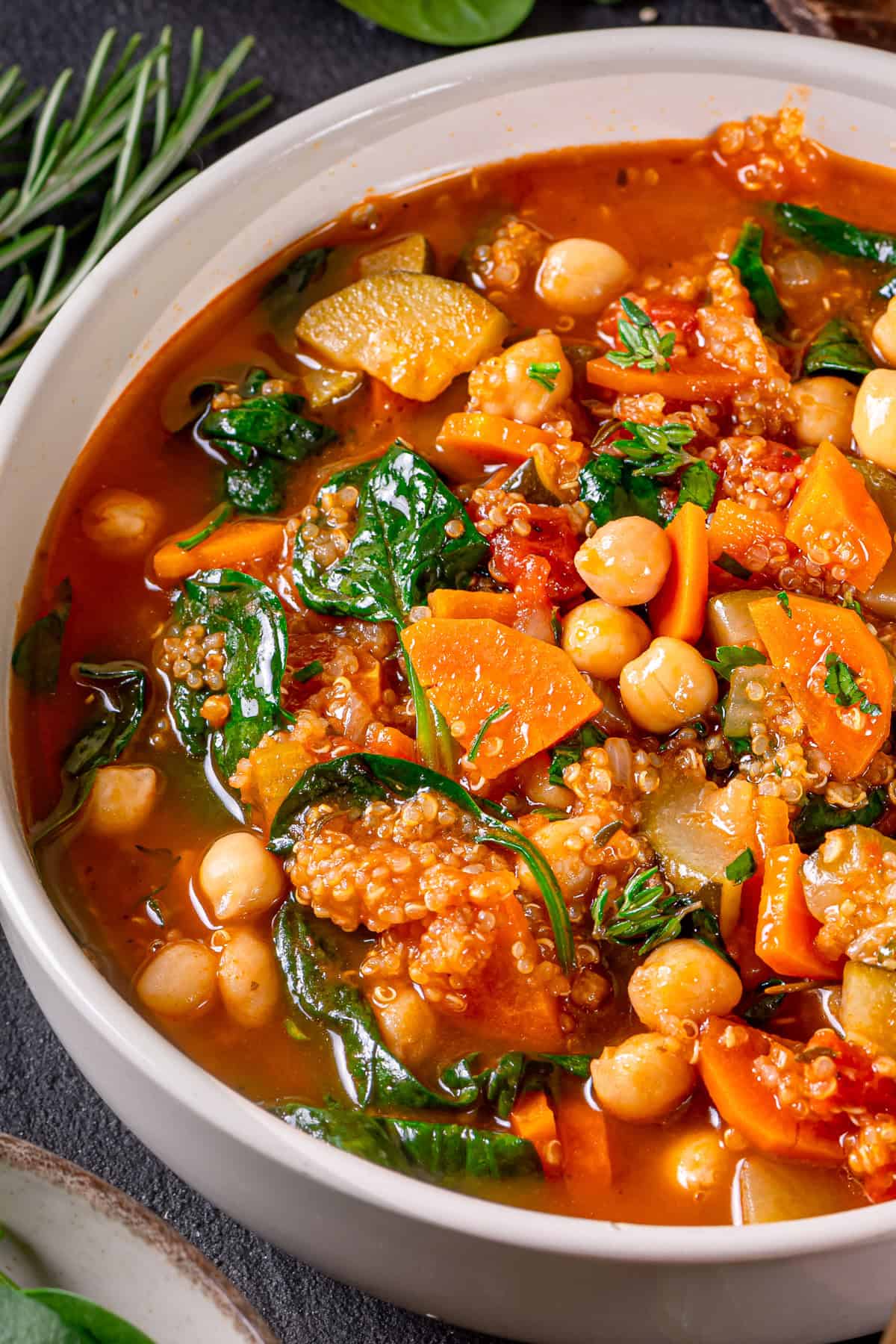 A close-up of a bowl filled with hearty vegetable quinoa soup featuring chickpeas, spinach, carrots, zucchini, quinoa, and a rich tomato-based broth, garnished with fresh herbs.