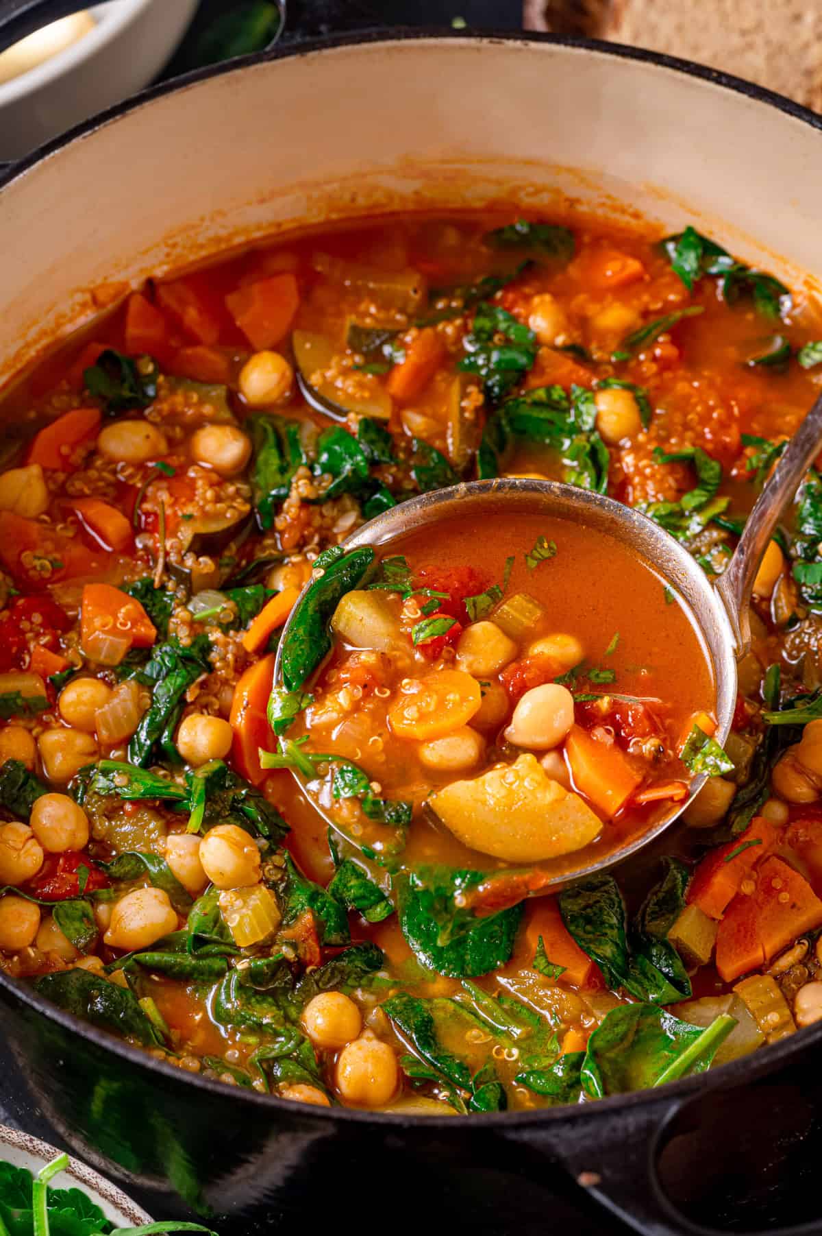 A ladle scoops up a hearty vegetable quinoa soup from a pot, filled with spinach, carrots, tomatoes, and herbs in a rich, colorful broth.