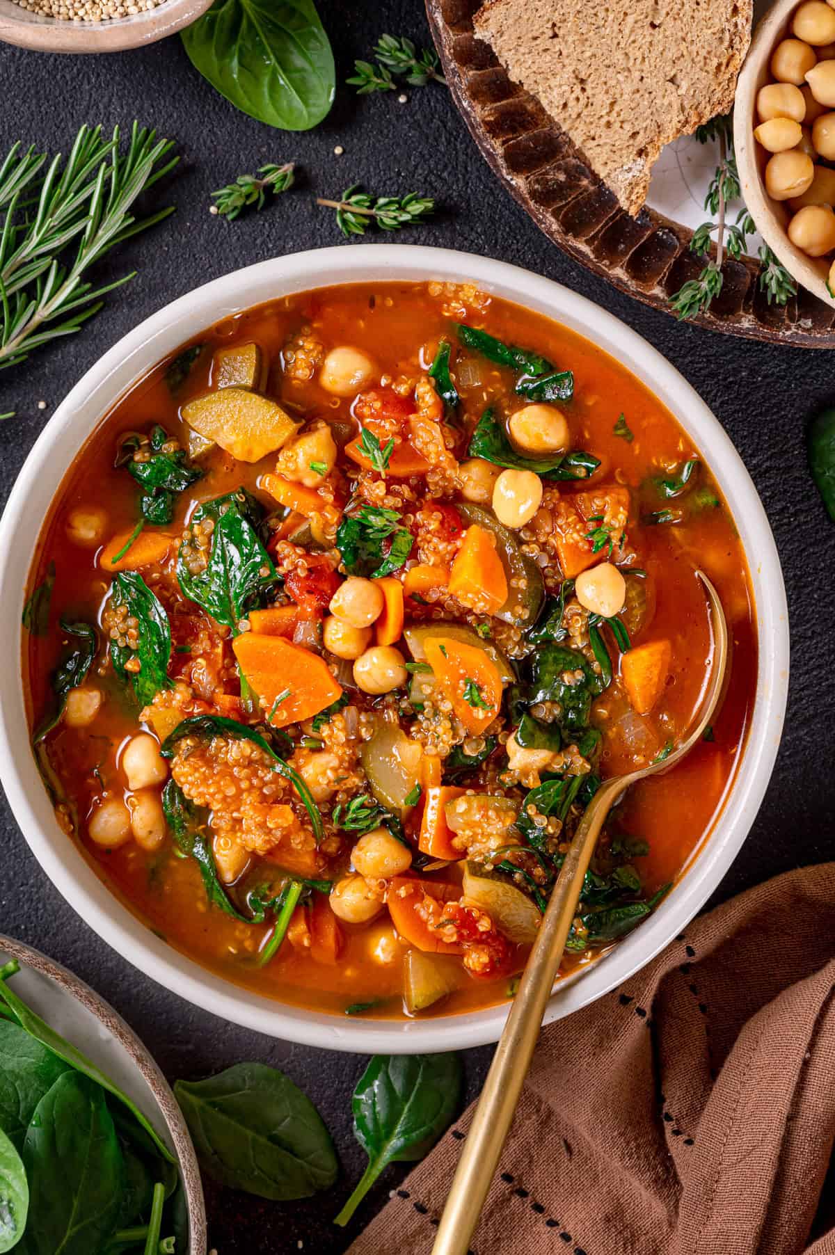 A bowl of vegetable quinoa soup with chickpeas, quinoa, spinach, carrots, and zucchini in a tomato broth, garnished with fresh herbs and surrounded by bread and spinach leaves on a dark surface.