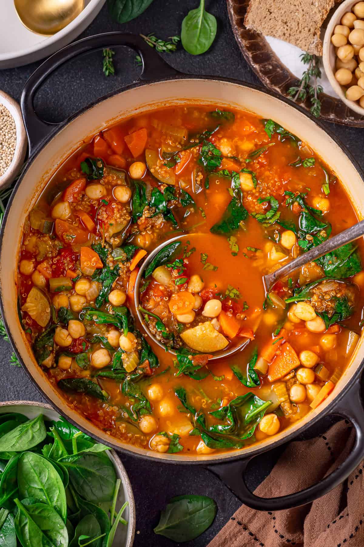 A pot of colorful vegetable quinoa soup with spinach, carrots, and tomatoes, surrounded by fresh spinach leaves, bread, and bowls of chickpeas and sesame seeds on a dark surface. A ladle rests in the stew.