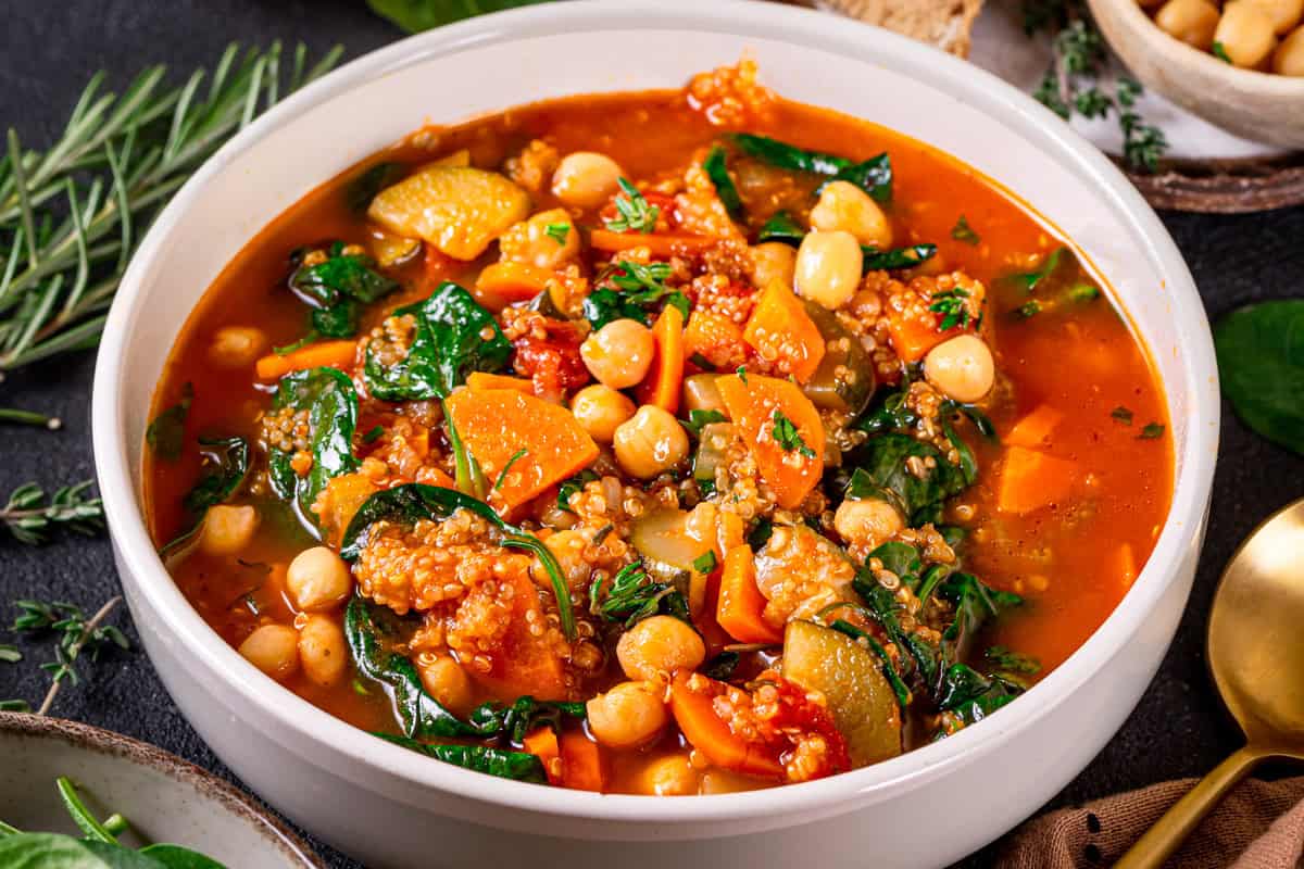 A close-up of a bowl of vegetable quinoa soup with chickpeas, carrots, spinach, tomatoes, and quinoa in a rich, red broth, garnished with fresh herbs. 