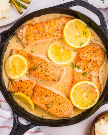 A skillet of creamy lemon dill salmon features four seared fillets, garnished with lemon slices and herbs. Sides of mashed potatoes, asparagus, and red onion are partially visible around the skillet.