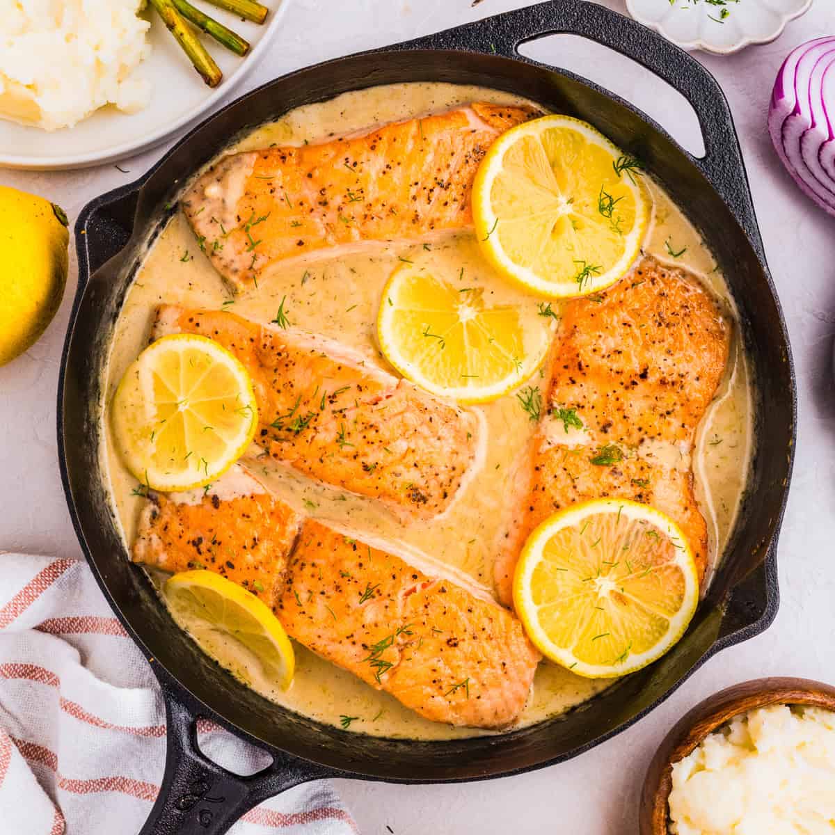 A skillet of creamy lemon dill salmon features four seared fillets, garnished with lemon slices and herbs. Sides of mashed potatoes, asparagus, and red onion are partially visible around the skillet.