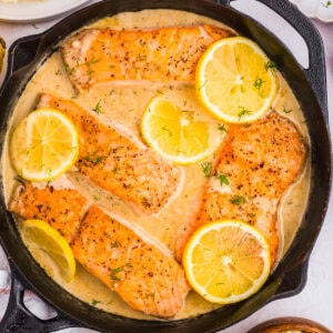 Lemon dill salmon fillets cooked in a creamy sauce, garnished with fresh dill and slices of lemon, served in a black skillet.