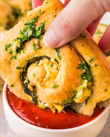 A hand dips a cheesy, herb-topped Spinach-Artichoke bread roll filled with spinach into a small bowl of red marinara sauce.