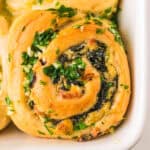 A close-up of a golden-brown, spiral Spinach Artichoke roll filled with spinach, cheese, and topped with fresh chopped parsley and garlic, resting in a white baking dish.