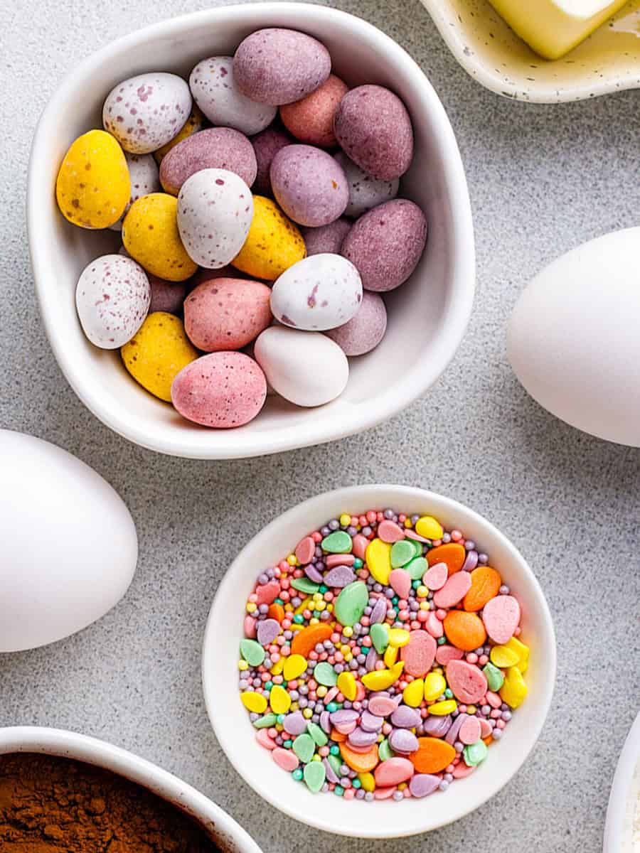 Two small bowls, one filled with pastel-colored speckled chocolate eggs and the other with assorted colorful sugar sprinkles, are surrounded by white eggs and ingredients on a light countertop.
