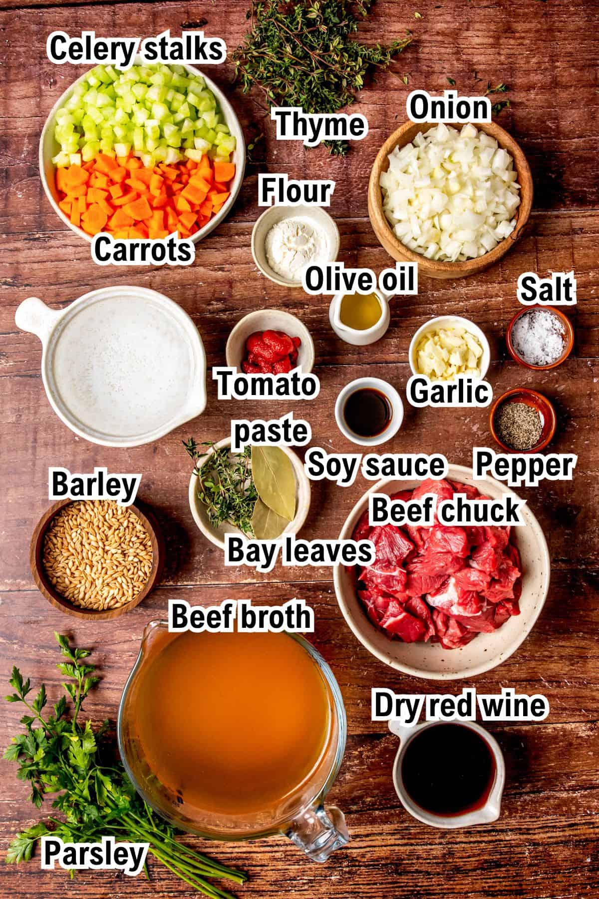 Top-down view of labeled ingredients for beef barley soup, including chopped celery, carrots, onion, garlic, raw beef chuck, beef broth, barley, tomato paste, flour, olive oil, dry red wine, soy sauce, herbs, salt, and pepper.