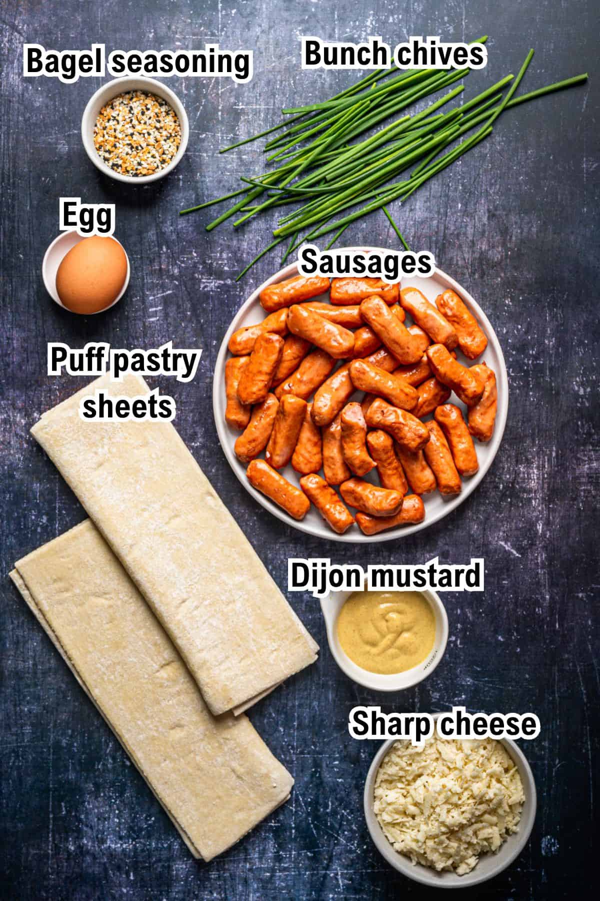 A flat lay of ingredients for Pigs in a Blanket on a dark surface: a bowl of bagel seasoning, a bunch of chives, a whole egg, a plate of small sausages, two puff pastry sheets, a bowl of Dijon mustard, and a bowl of shredded sharp cheese.