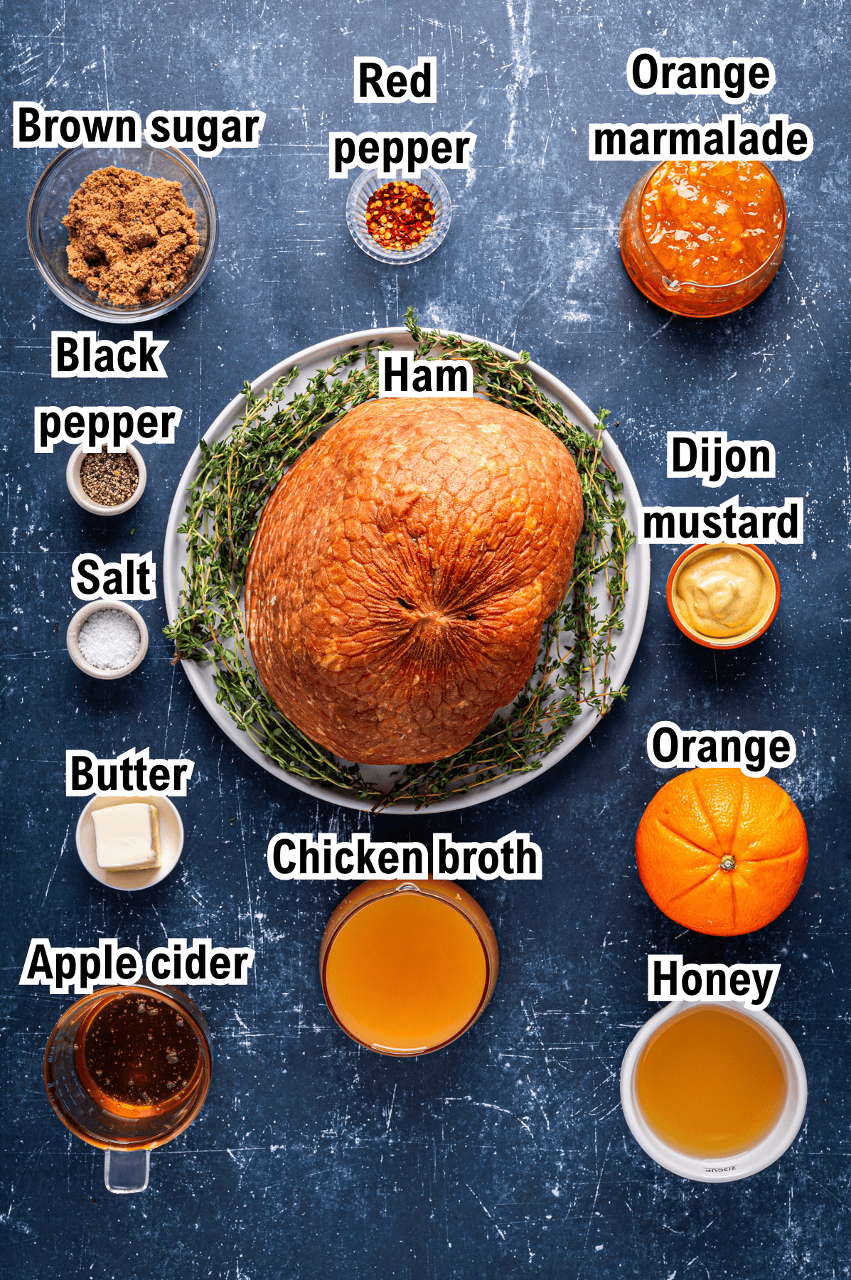 A large ham on a plate surrounded by labeled ingredients for easy ham glaze: brown sugar, red pepper, orange marmalade, black pepper, salt, Dijon mustard, butter, chicken broth, orange, apple cider, and honey on a blue surface.
