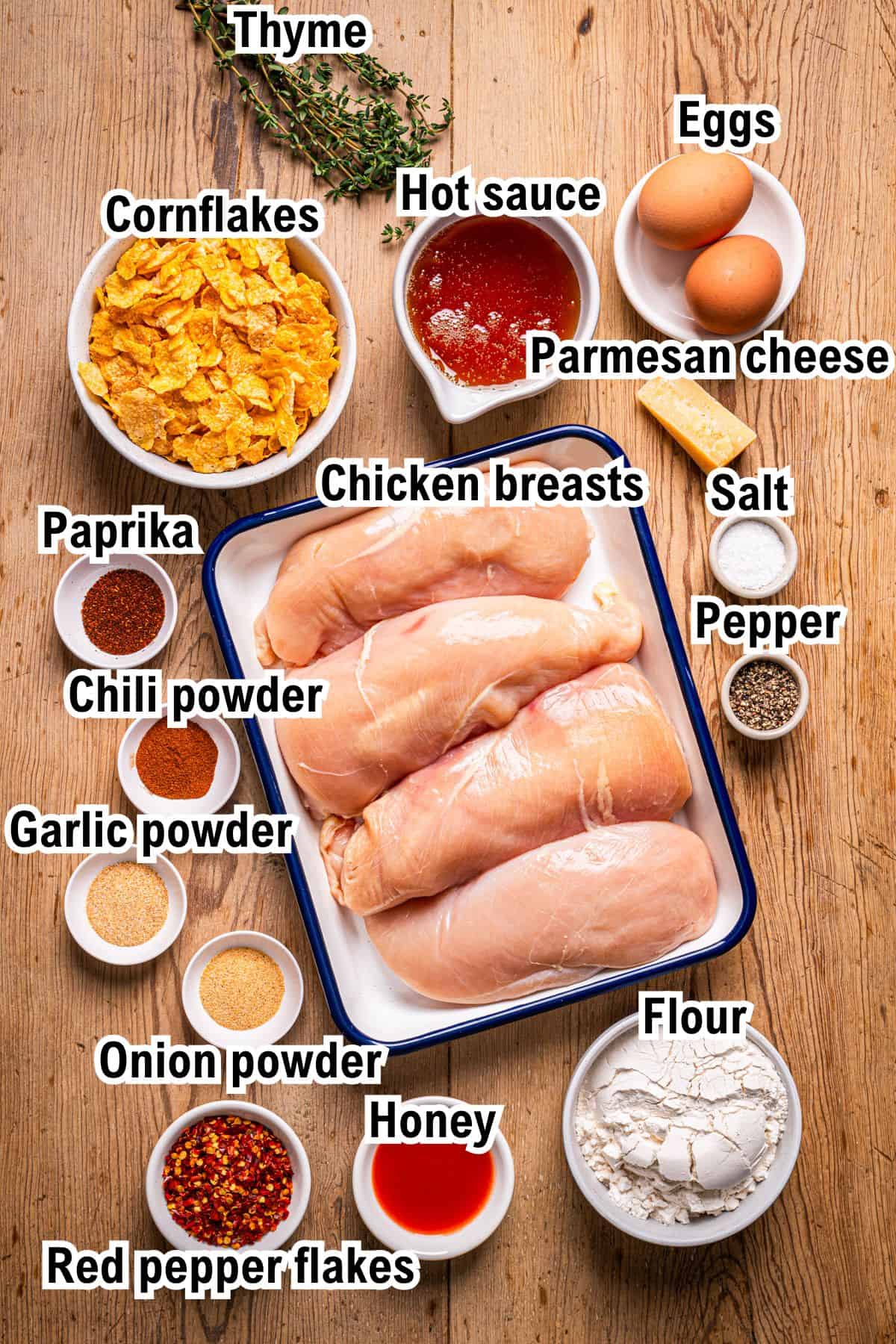 The Ingredients for Crispy Baked Hot Honey Chicken A wooden table with labeled ingredients for a hot honey baked chicken recipe, including chicken breasts, eggs, flour, cornflakes, Parmesan cheese, hot sauce, honey, spices (paprika, chili, onion, garlic powder), salt, pepper, red pepper flakes, and thyme.