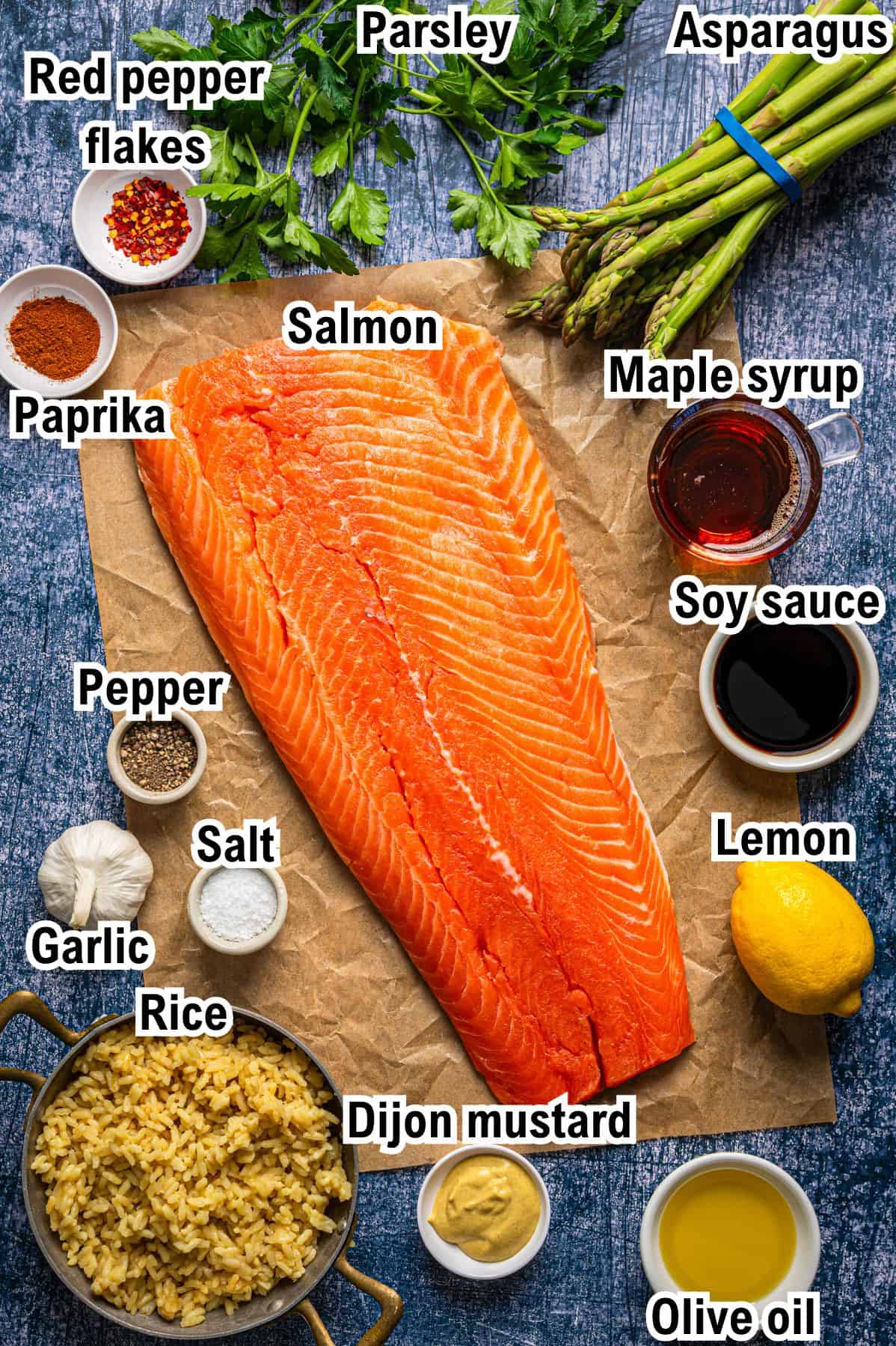 Overhead view of raw salmon fillet on parchment paper, surrounded by labeled ingredients: asparagus, parsley, maple syrup, soy sauce, lemon, olive oil, Dijon mustard, rice, garlic, salt, pepper, paprika, and red pepper flakes.