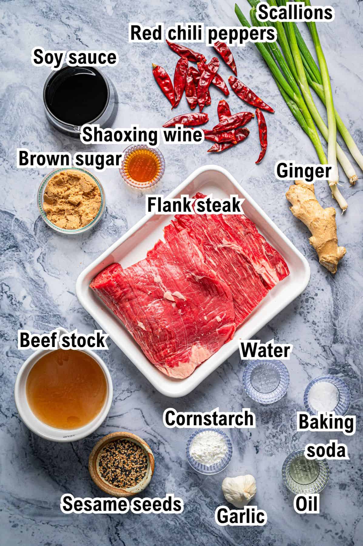 A marble surface displays labeled ingredients for a Mongolian Beef recipe: soy sauce, Shaoxing wine, brown sugar, flank steak, beef stock, sesame seeds, red chili peppers, scallions, ginger, cornstarch, water, garlic, baking soda, and oil.