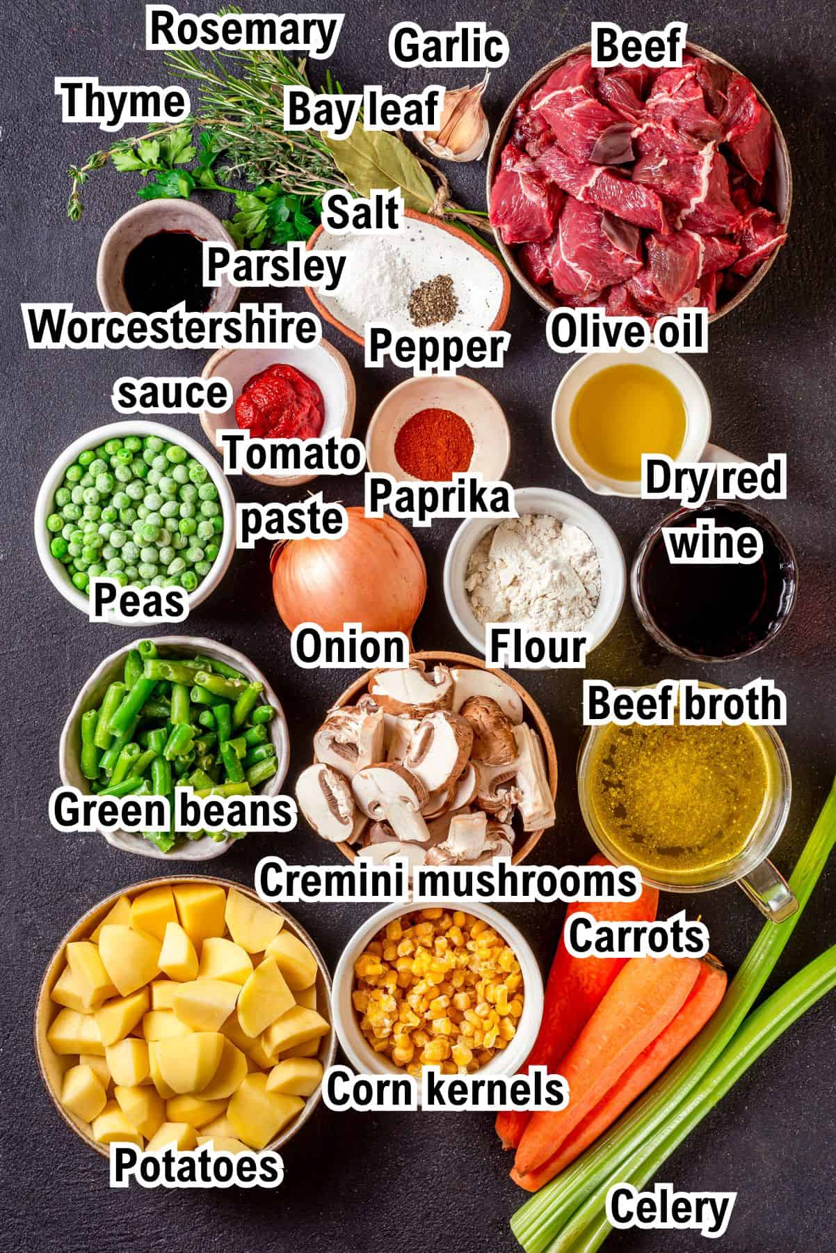 The Ingredients for Mulligan Stew (Hobo Stew) Top-down view of labeled ingredients for Mulligan beef stew, including beef, olive oil, dry red wine, beef broth, carrots, celery, potatoes, corn, peas, green beans, mushrooms, onion, tomato paste, flour, spices, and herbs.