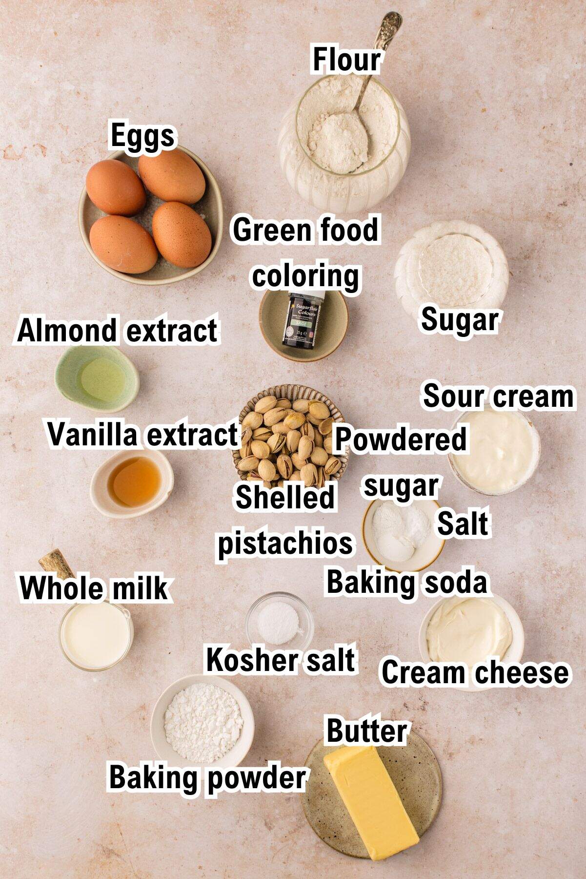 A flat lay of labeled baking ingredients for Pistachio Bundt Cake in bowls including eggs, flour, sugar, sour cream, baking powder, baking soda, kosher salt, salt, cream cheese, butter, whole milk, vanilla extract, almond extract, powdered sugar, green food coloring, and shelled pistachios.