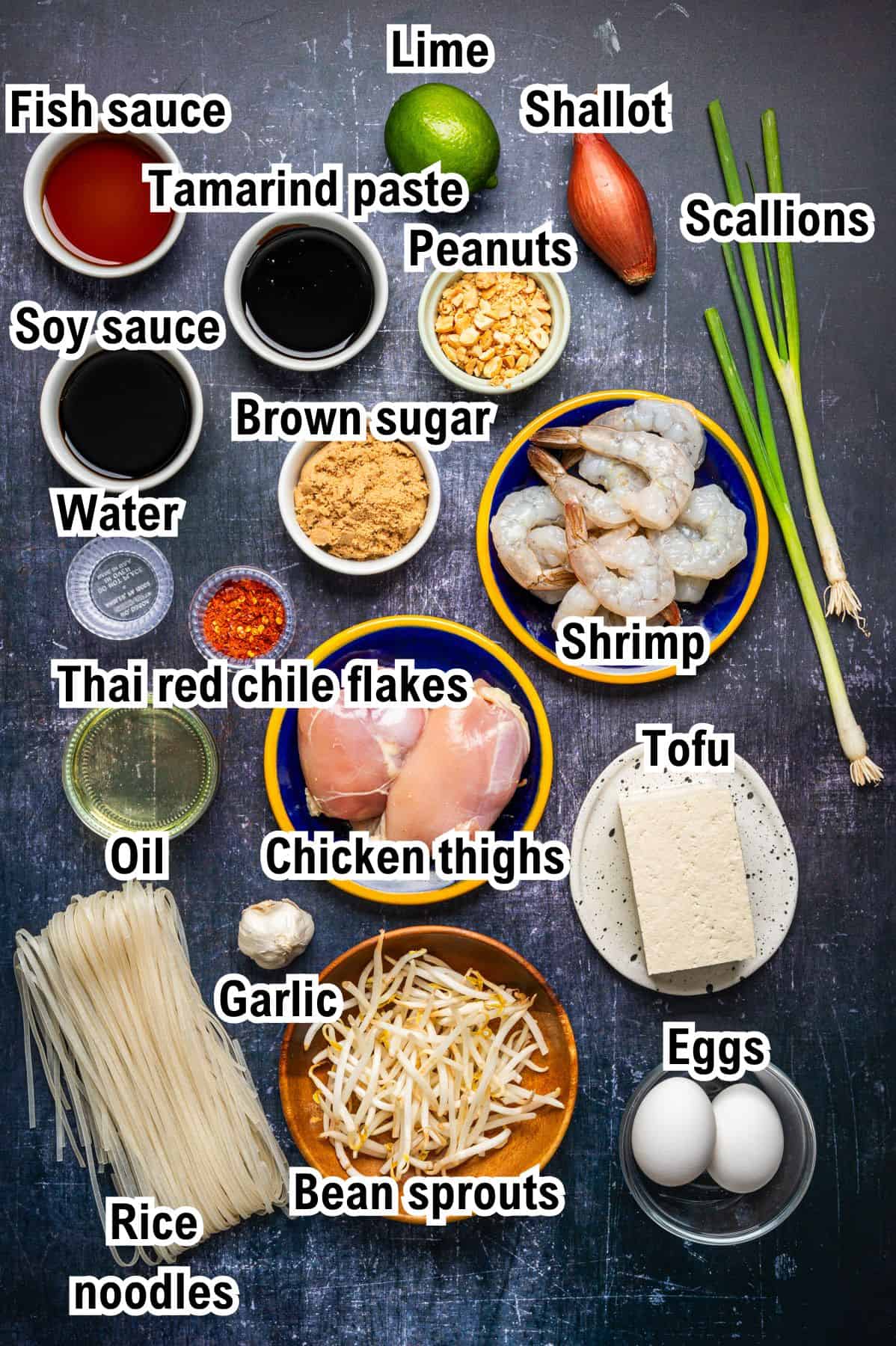 Top-down view of Pad Thai ingredients on a dark surface, labeled with text: lime, shallot, scallions, peanuts, shrimp, tofu, eggs, chicken thighs, garlic, bean sprouts, rice noodles, oil, Thai red chile flakes, brown sugar, soy sauce, tamarind paste, fish sauce, and water.