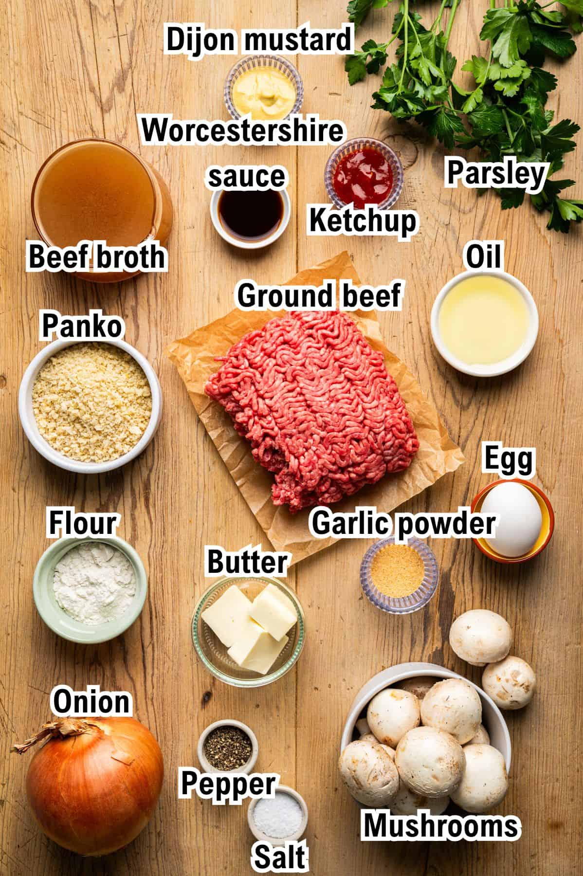 A wooden table displays ground beef surrounded by labeled ingredients: Dijon mustard, Worcestershire sauce, ketchup, parsley, oil, egg, garlic powder, mushrooms, salt, pepper, onion, butter, flour, panko, and beef broth.