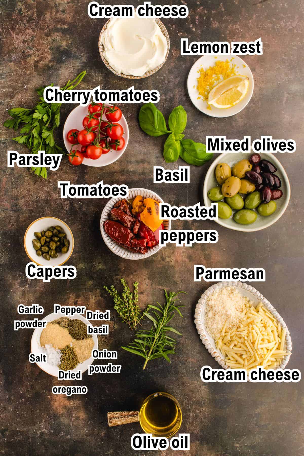 A variety of labeled ingredients for a Mediterranean cheese log are arranged on a dark surface, including cream cheese, cherry tomatoes, lemon zest, olives, parmesan, spices, capers, parsley, basil, roasted peppers, and olive oil.