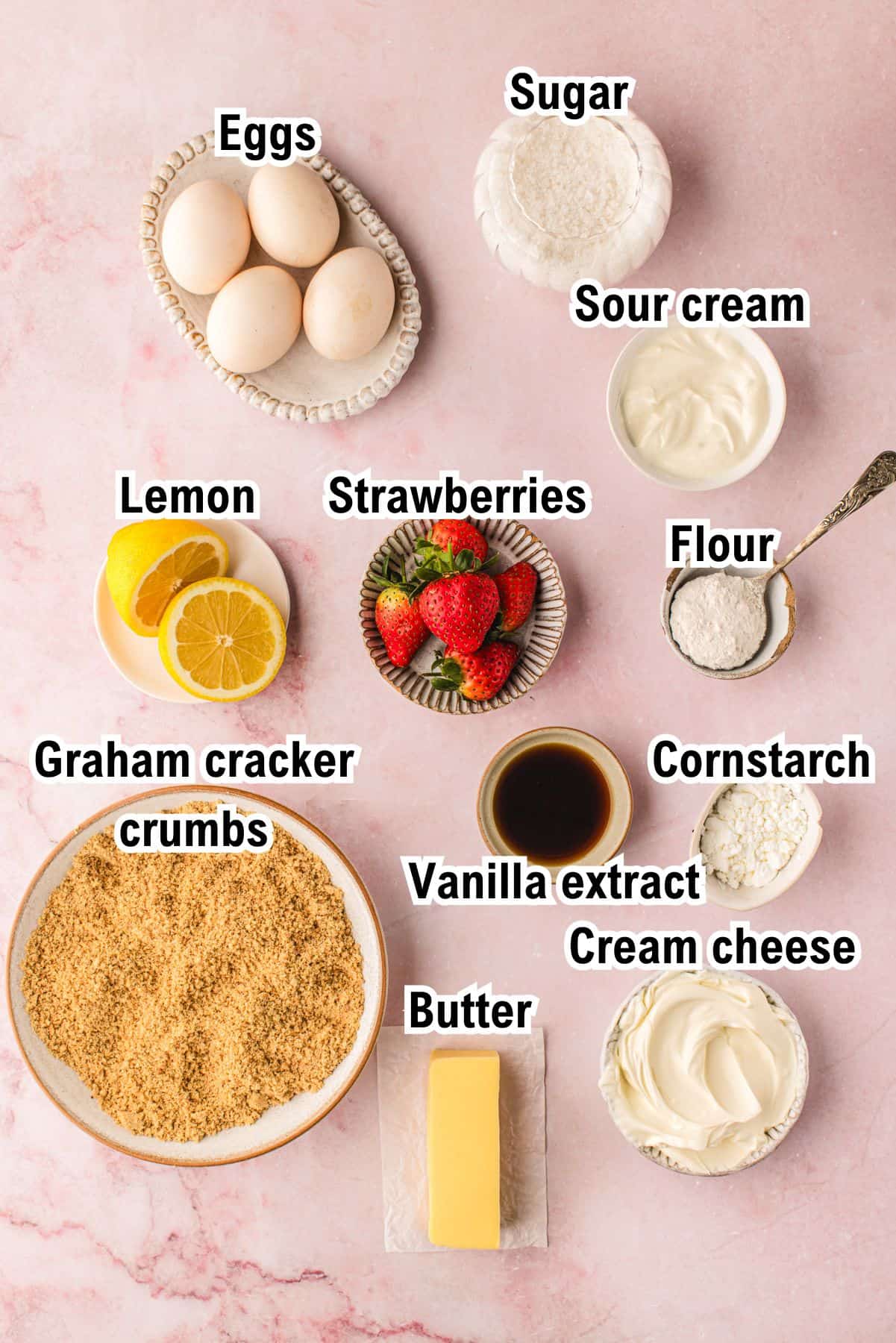 The Ingredients for Classic New York-Style Cheesecake This photo shows the ingredients for Classic New York-Style Cheesecake: A flat lay of labeled baking ingredients: eggs, sugar, sour cream, lemon, strawberries, flour, cornstarch, graham cracker crumbs, vanilla extract, cream cheese, and a stick of butter.