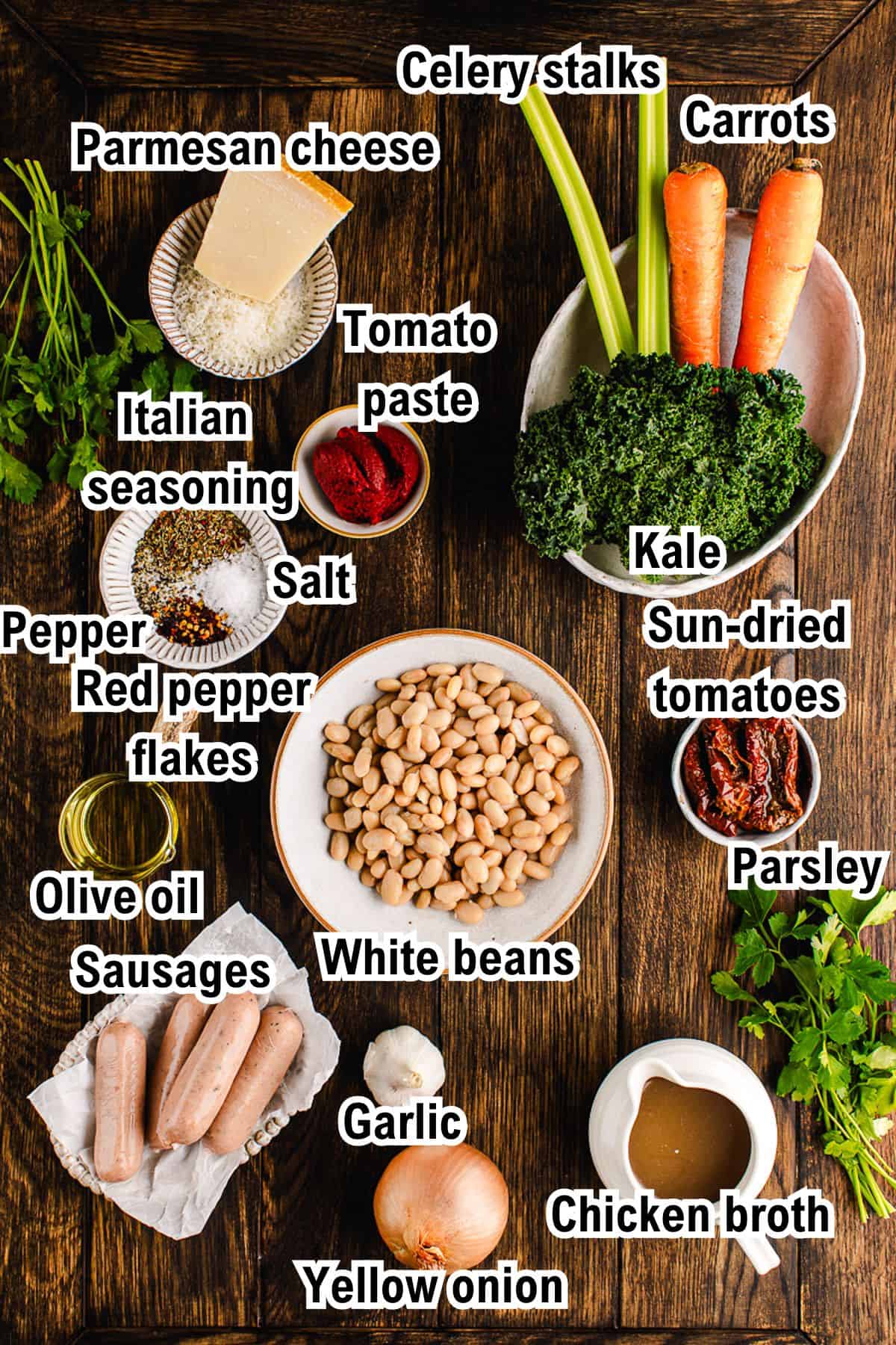 The Ingredients for Tuscan White Bean Soup A flat lay of Tuscan White Bean Soup ingredients on a wooden table, including carrots, celery, kale, parsley, Parmesan cheese, Italian seasoning, tomato paste, sun-dried tomatoes, sausages, white beans, garlic, yellow onion, chicken broth, olive oil, salt, pepper, and red pepper flakes.