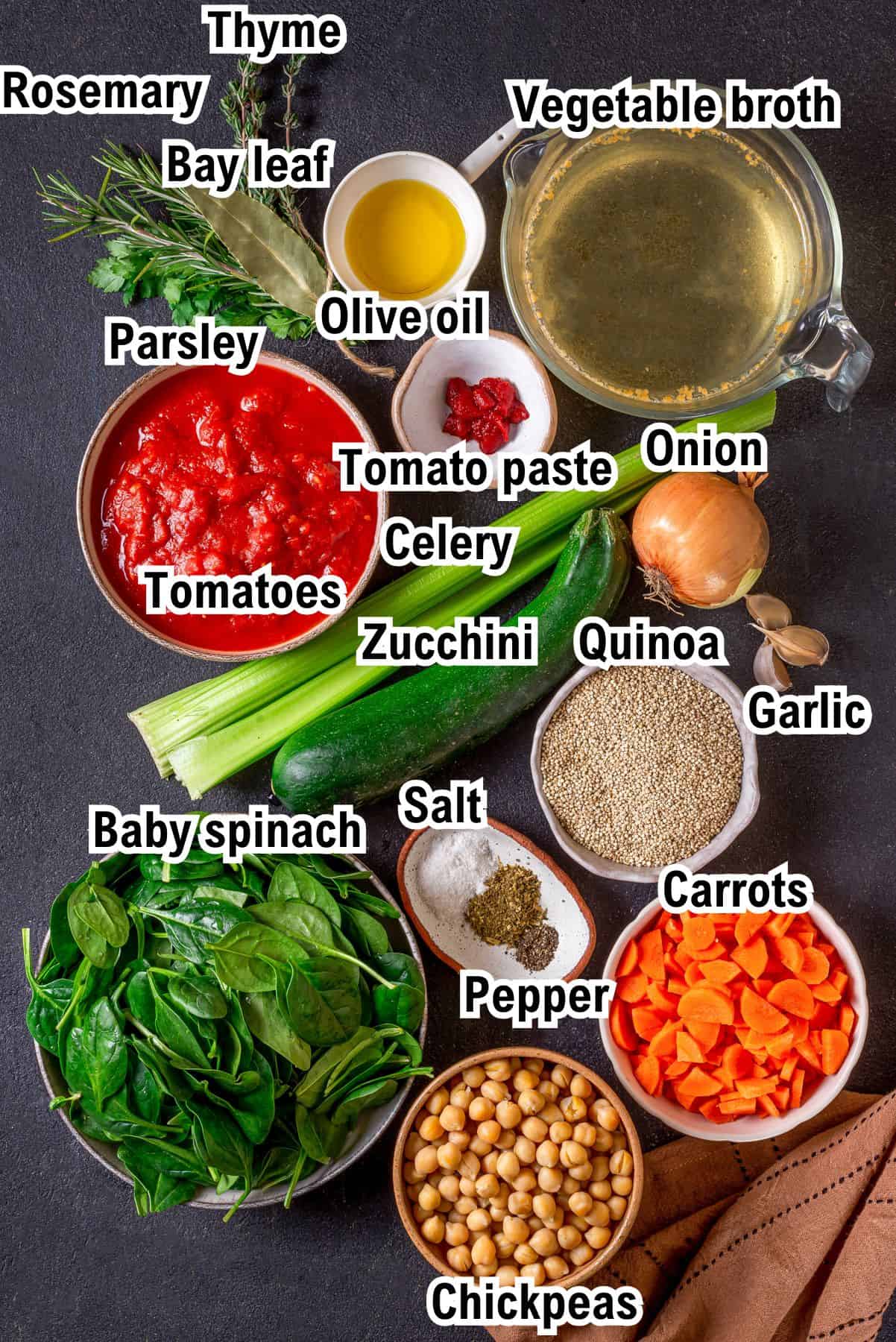 A flat lay of fresh vegetable quinoa soup ingredients including tomatoes, carrots, onion, celery, zucchini, spinach, garlic, parsley, thyme, bay leaf, quinoa, chickpeas, salt, pepper, olive oil, and vegetable broth.